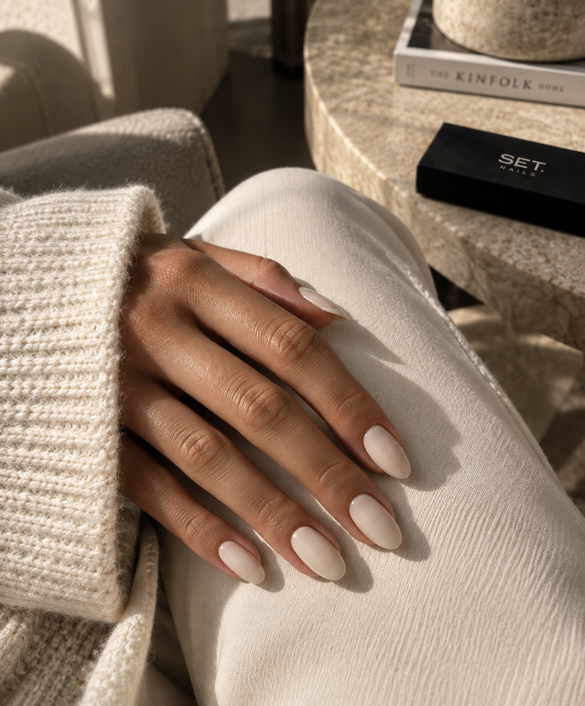 Hand resting on ivory fabric wearing SET. 002 BONE warm neutral almond nails in soft daylight.